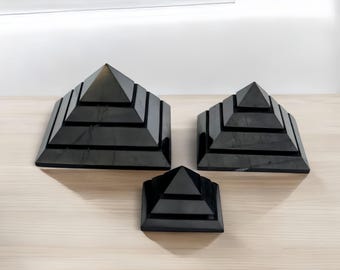 May include: Three black pyramid-shaped objects of varying sizes, made of polished stone, arranged on a light wood surface.