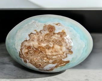 May include: A large, oval-shaped stone with a smooth, polished surface. The stone features a blend of colors, including light blue, white, and brown. The brown areas have a textured, earthy appearance, while the blue and white areas are more smooth.