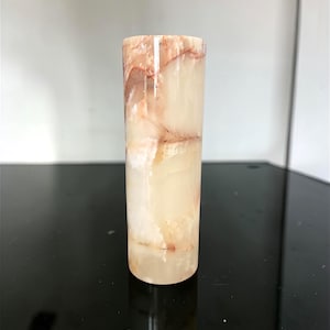 May include: A cylindrical, translucent stone vase with a polished finish. The vase is primarily cream-colored with streaks of reddish-brown and tan throughout. The vase is standing upright.