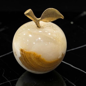 May include: A decorative apple sculpture crafted from polished cream and white stone with distinct brown veining. The fruit features a smooth, rounded body topped with a textured, metallic gold-toned stem and a single leaf extending to the right side.