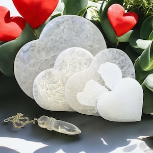 May include: Three white selenite heart-shaped crystals with etched angel wing designs. The crystals are arranged in a nested pattern, with the largest heart at the back and the smallest heart in front. A clear quartz pendulum is lying on the table in front of the crystals.