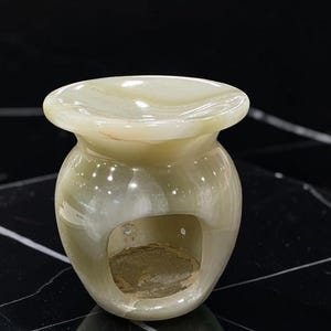 May include: A pale green stone oil burner with a smooth, polished finish. The vessel features a wide, circular top basin for oil and a hollowed-out arched opening at the base designed to accommodate a small heat source for warming the contents.