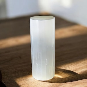 Peut inclure: Une tour de cristal de sélénite blanche, également appelée baguette de sélénite, est une forme cylindrique lisse avec une apparence blanche translucide. Le cristal est posé sur une surface en bois.