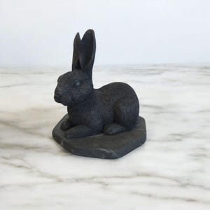Puede incluir: Una figurita de conejo negro sentada sobre una base de piedra negra. El conejo está esculpido en un estilo realista con pelo detallado y una oreja larga y caída.