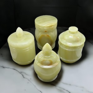 Puede incluir: Conjunto de cuatro jarrones decorativos hechos de ónix verde. Los jarrones tienen un acabado liso y pulido y están tallados intrincadamente con un diseño único. Cada jarrón tiene una tapa con un pomo en la parte superior.