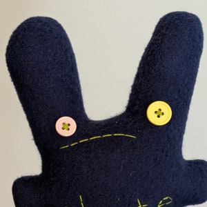 May include: A handmade navy blue felt bunny toy with button eyes and a stitched mouth. The bunny has a yellow button on one side and a pink button on the other. The mouth is stitched with yellow thread.