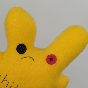 May include: A yellow felt plush toy shaped like a star with a black button for an eye and a pink button on the side. The toy has a drawn-on frown and the word "chit" written on the bottom.