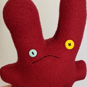 May include: A red felt plush toy with two long arms and a sad face. The toy has one blue button eye and one yellow button eye with a black X in the center.
