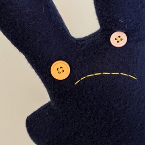 May include: A dark blue felt toy with a sad face. The face has two buttons for eyes, one orange and one pink, and a yellow stitched mouth.