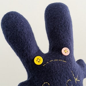 May include: A dark blue felt hand puppet with two yellow buttons and one pink button. The puppet has a stitched smile and a stitched arrow on the right side.