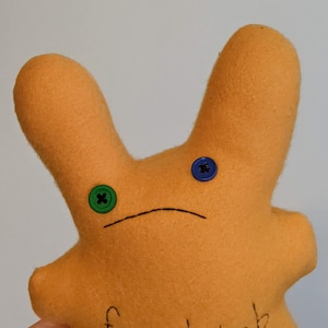 May include: Orange plush toy with button eyes and a stitched mouth. The toy has long, upright ears and a rounded body. A green button is on the left side and a blue button is on the right side. The toy is handmade.