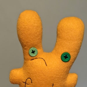 May include: A plush orange toy with two green buttons for eyes. The toy has a stitched line across the face and a stitched line down the center of the body.