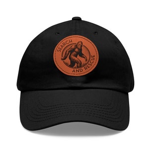 以下が含まれることがあります： ビッグフットのシルエットと「Search and Rescue」の文字が書かれた茶色の革のパッチが付いた黒いベースボールキャップ。
