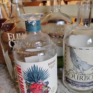 Puede incluir: Varias botellas de licor vacías de vidrio, incluyendo una de tequila con una etiqueta colorida con un corazón y una planta de agave, y una de bourbon con una ilustración de ballena. Se ven diferentes formas y tamaños.