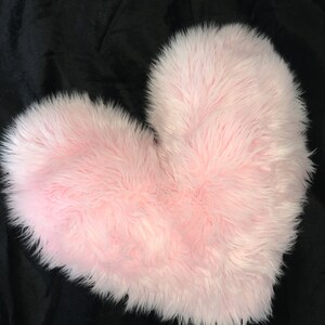 May include: A fluffy, pink heart-shaped pillow. The pillow has a soft, faux fur texture and is set against a black background. The heart shape is clearly defined, with a rounded top and a pointed bottom.