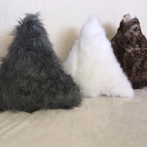 May include: Three triangular-shaped decorative pillows in a row. The pillows are covered in faux fur. The colors are dark gray, white, and brown with a speckled pattern. The pillows are arranged on a light beige surface.