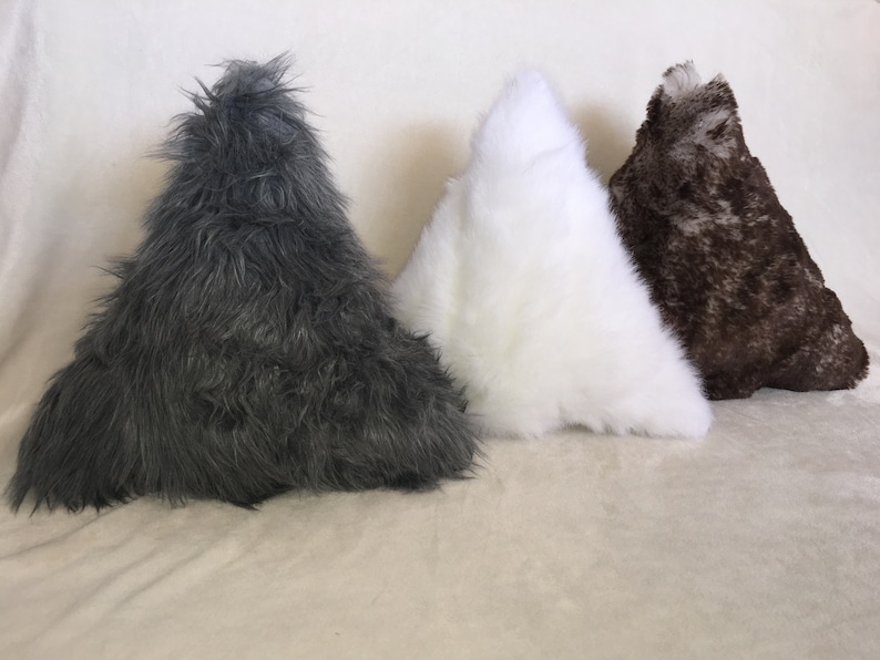 May include: Three triangular-shaped decorative pillows in a row. The pillows are covered in faux fur. The colors are dark gray, white, and brown with a speckled pattern. The pillows are arranged on a light beige surface.