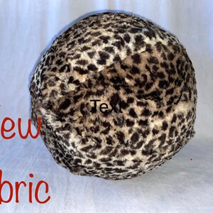 May include: A round, plush item with a leopard print pattern in shades of brown, tan, and black. The words "New fabric" are written in red. The item appears to be made of soft, faux fur.