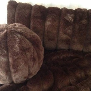 May include: A brown faux fur pillow and throw set. The pillow is round and has a segmented design. The throw and the back of the chair are covered in the same brown faux fur material. The fur has a soft, plush appearance.