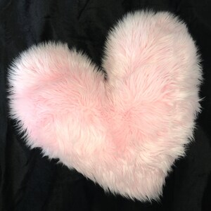 May include: A fluffy, light pink heart-shaped pillow. The pillow has a soft, furry texture and is set against a black background. The heart shape is clearly defined, making it a decorative item.