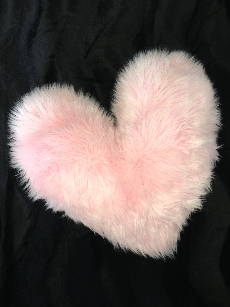 May include: A fluffy, light pink heart-shaped pillow. The pillow has a soft, furry texture and is set against a black background. The heart shape is clearly defined, making it a decorative item.