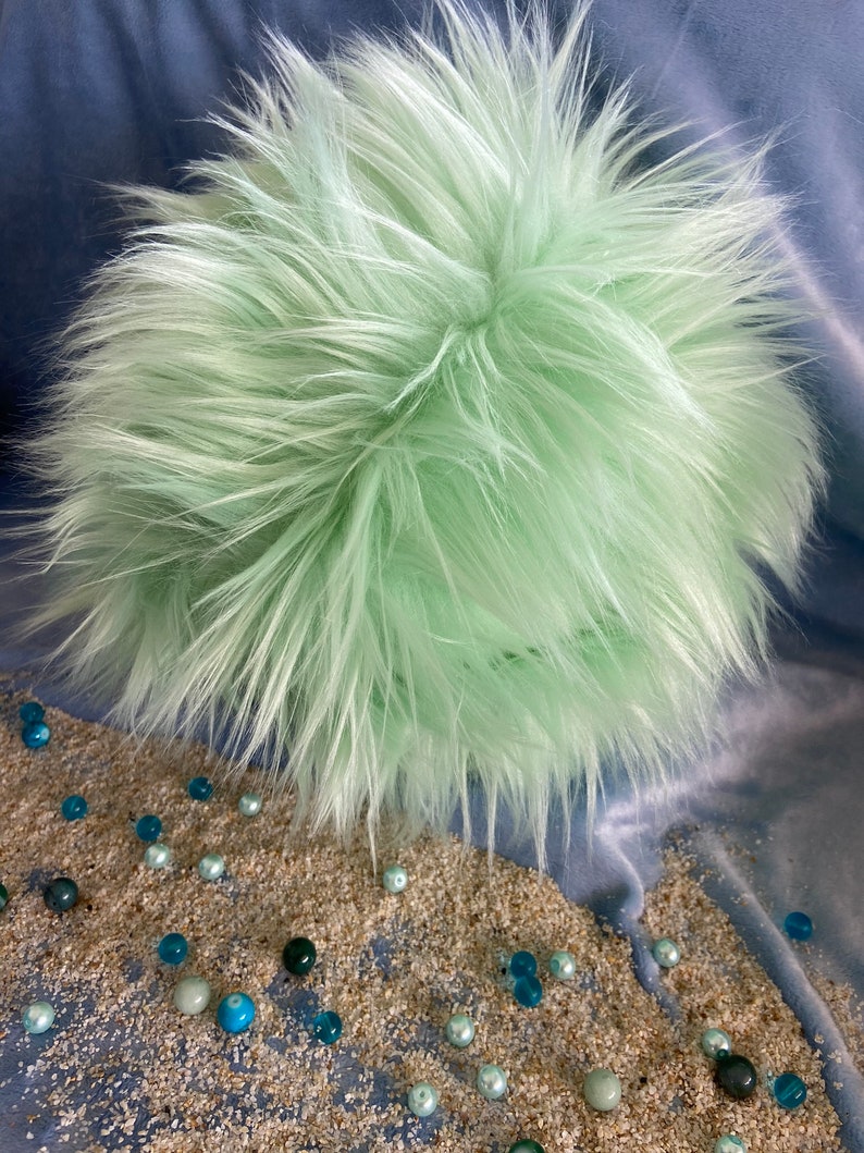 May include: A light green fluffy ball with a soft, plush texture. The ball is sitting on a surface covered in small, blue and white beads.