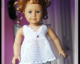 May include: A doll wearing a white crocheted dress with a pink trim and matching crocheted pants. The doll has long, curly red hair and blue eyes.