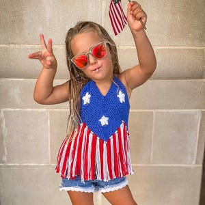 May include: A young child wearing a patriotic outfit, including a blue and white top with red and white fringe, denim shorts, and red sunglasses. The child is holding a small American flag.