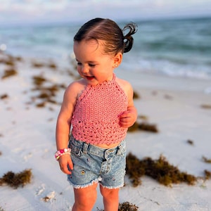 Puede incluir: Una niña pequeña con un top halter rosa de ganchillo y pantalones cortos de mezclilla con detalles bordados. La niña también lleva una pulsera con cuentas rosas y blancas. La niña está en una playa de arena.