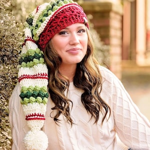 Puede incluir: Un gorro de elfo de crochet blanco, rojo y verde con un pompón. El gorro tiene una cola larga y puntiaguda con un pompón blanco en el extremo.