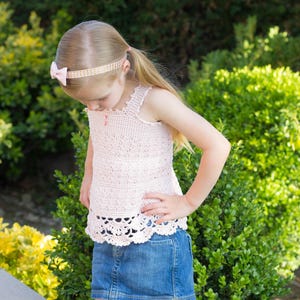 May include: A pale pink, sleeveless, crocheted top with a scalloped hem is worn with a denim skirt. The outfit is completed with a pink headband and pink sandals. The ensemble is suitable for a child.