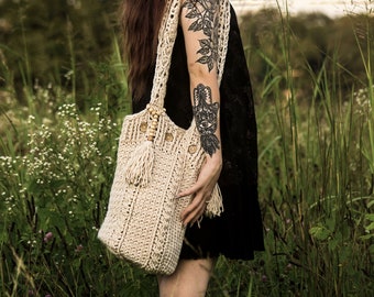 May include: A woman with long dark hair and tattoos is standing in a field of tall grass. She is wearing a black dress and a crocheted tote bag with wooden beads and tassels.