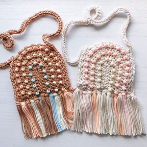 May include: Two handmade crochet bags with beaded details and fringe. One is brown with peach, blue, and cream beads and fringe. The other is off-white with pink beads and fringe. Both have long straps.