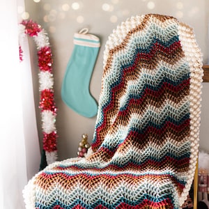 May include: A handmade, colorful crochet blanket draped over a wooden chair. The blanket features a chevron pattern in shades of red, brown, teal, and cream. The edges are finished with a textured, bobble stitch border. The chair has dark wooden legs.
