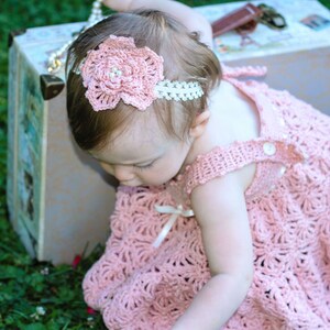 May include: A baby girl wearing a pink crocheted dress with a white crocheted flower headband. The baby is sitting in front of a vintage suitcase.