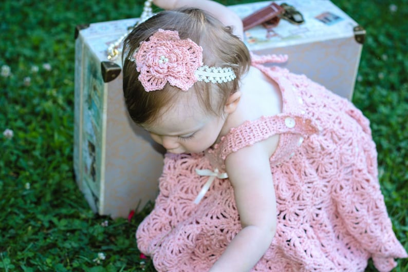 May include: A baby girl wearing a pink crocheted dress with a white crocheted flower headband. The baby is sitting in front of a vintage suitcase.