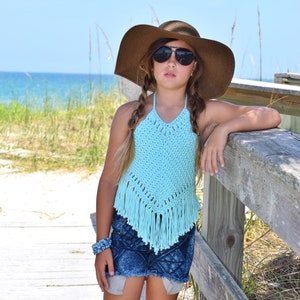 Puede incluir: Una persona joven lleva un top halter de crochet azul claro con flecos, un sombrero marrón de ala ancha y pantalones cortos de mezclilla. Está de pie en una pasarela de madera con una playa en el fondo.