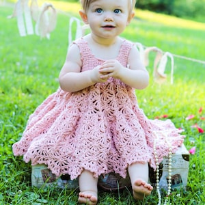 May include: A young child wearing a pink crocheted dress with a flower headband sits on a vintage suitcase with a pearl necklace draped over it. The child is sitting in a grassy area with green foliage.