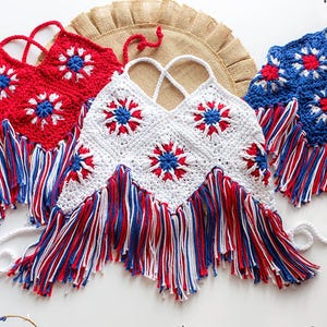May include: Handmade crochet tops in red, white, and blue, featuring starburst designs and fringe. The tops have halter straps and are arranged on a white surface with a burlap accent, perfect for patriotic celebrations.