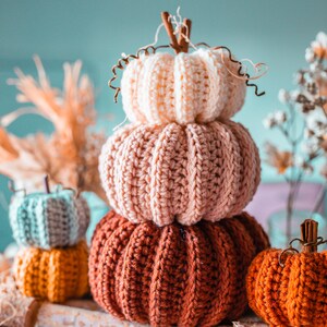 May include: A stack of three crocheted pumpkins in shades of orange, pink, and cream, with a small orange pumpkin and two smaller pumpkins in the background. The pumpkins have brown stems and are arranged on a wooden surface.