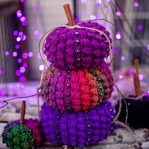 Puede incluir: Tres calabazas de ganchillo en tonos morados, rosas y naranjas, decoradas con cuentas y palitos de canela. Las calabazas están apiladas sobre corteza de abedul. Luces moradas en el fondo crean un ambiente festivo.