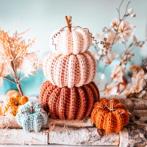 May include: A stack of three knitted pumpkins in shades of white, pink, and rust-orange, with a small orange pumpkin and a blue-green pumpkin in front. The pumpkins are arranged on a birch log, with fall foliage in the background.