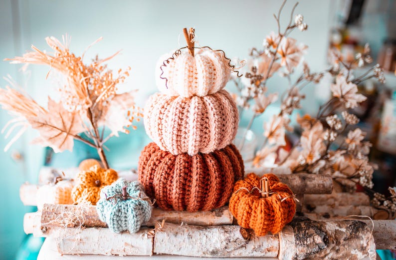 May include: A stack of three knitted pumpkins in shades of white, pink, and rust-orange, with a small orange pumpkin and a blue-green pumpkin in front. The pumpkins are arranged on a birch log, with fall foliage in the background.