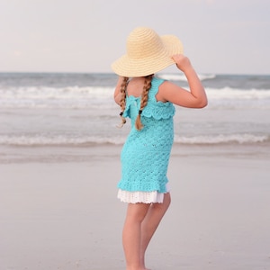 K&ouml;nnte beinhalten: Ein junges M&auml;dchen in einem hellblauen H&auml;kelkleid und einem breitkrempigen Strohhut steht an einem Sandstrand und blickt auf das Meer.