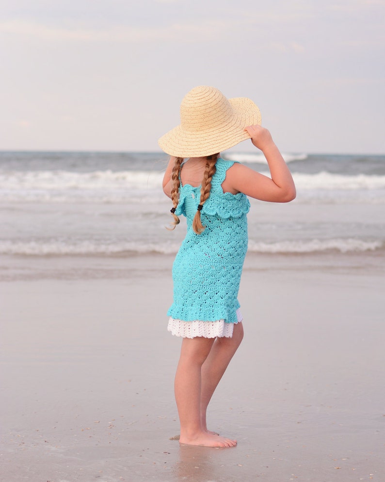 K&ouml;nnte beinhalten: Ein junges M&auml;dchen in einem hellblauen H&auml;kelkleid und einem breitkrempigen Strohhut steht an einem Sandstrand und blickt auf das Meer.