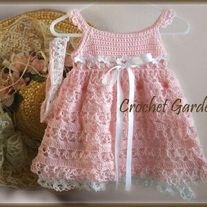 May include: A handmade pink crochet dress with white ribbon and lace trim, hanging on a white hanger. The dress features a delicate openwork design and the text "Crochet Garden". A straw hat with lace and flowers is in the background.