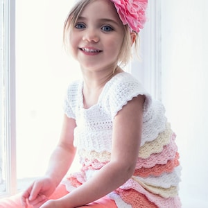 May include: A young girl wearing a white crocheted dress with multiple layers of ruffled trim in shades of pink, yellow, and white. She is wearing a pink flower headband and has a bright smile.