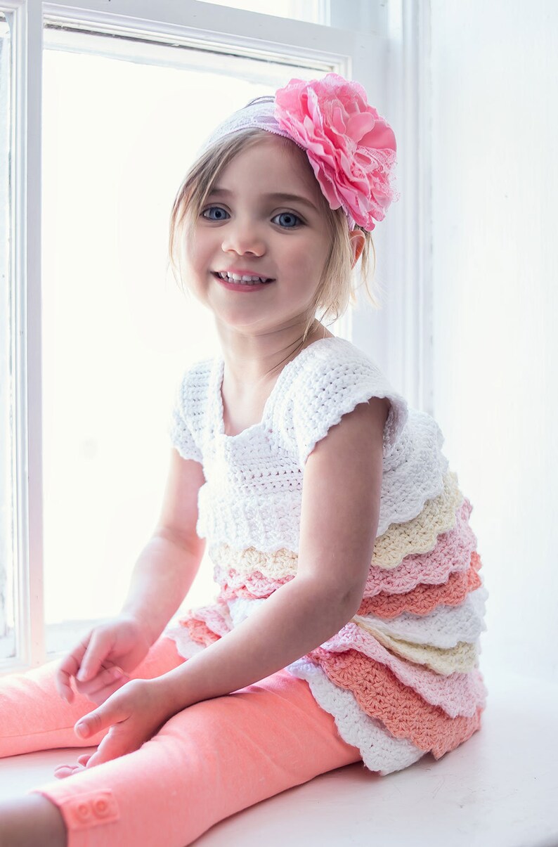 May include: A young girl wearing a white crocheted dress with multiple layers of ruffled trim in shades of pink, yellow, and white. She is wearing a pink flower headband and has a bright smile.