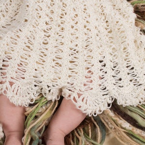 May include: A white crocheted baby blanket with a delicate, lacy pattern. The blanket is draped over a baby's legs, with the bottom edge of the blanket resting on a bed of soft, colorful yarn.