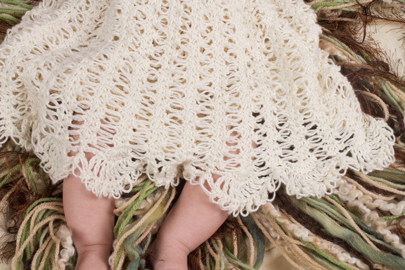 May include: A white crocheted baby blanket with a delicate, lacy pattern. The blanket is draped over a baby's legs, with the bottom edge of the blanket resting on a bed of soft, colorful yarn.